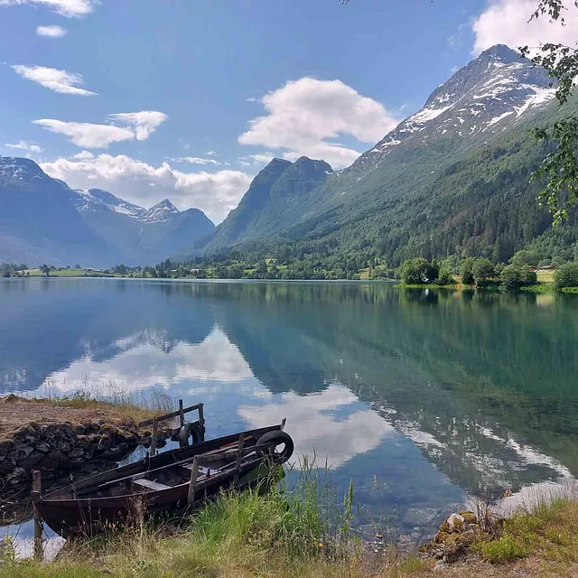 To gamle trebåter ligger i vannkanten ved en speilblank innsjø som reflekterer høye, grønne fjell.