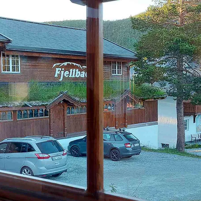 Utsikt gjennom et vindu mot en parkeringsplass foran et stort trebygg med torv på taket.