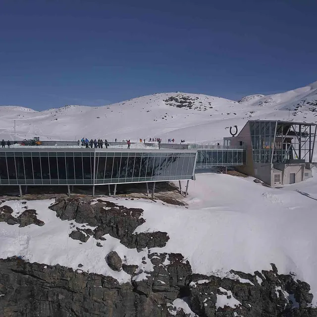 Moderne restaurantbygg i glass og betong, plassert på en snødekket fjellhøyde.