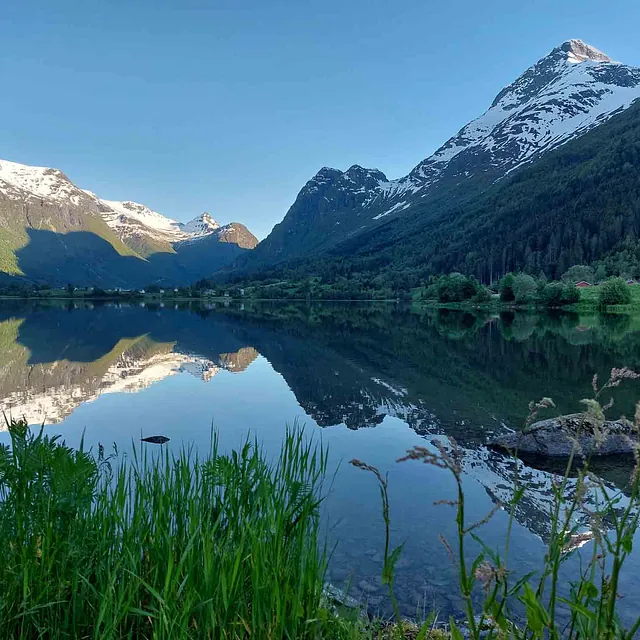 Grønt gress i forgrunnen ved en speilblank innsjø som reflekterer høye, delvis snødekte fjell.