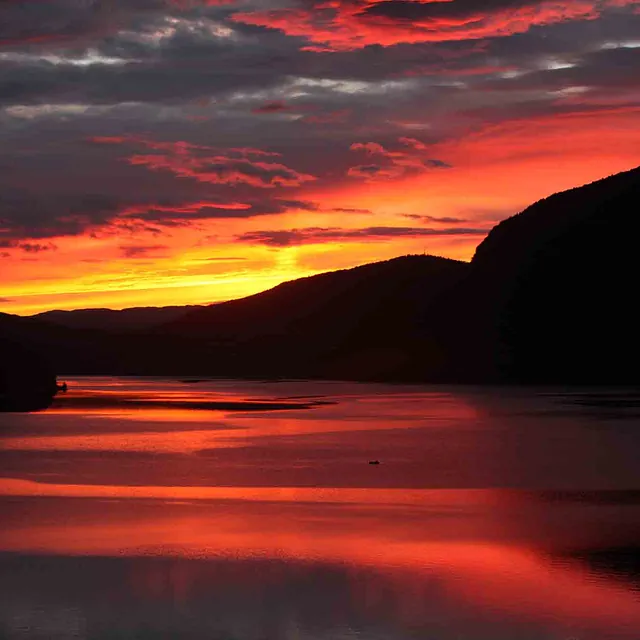 Fargesprakende, rød og oransje solnedgang over en mørk fjord og fjellside.