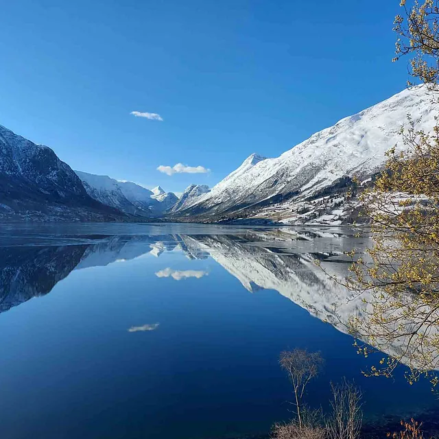 Speilblank, mørk fjord som reflekterer snødekte fjell og en skyfri, blå himmel.