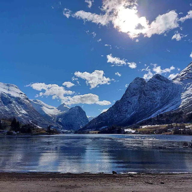 Utsikt over en delvis islagt innsjø omkranset av bratte, snødekte fjellsider under en blå himmel med sol.