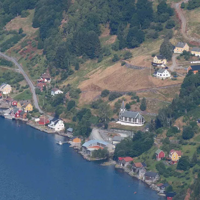 Flyfoto av en bratt fjellside som skråner ned mot fjorden, med en bilvei og spredt bebyggelse.