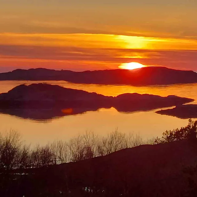Fargesprakende, dyp oransje solnedgang over et kystlandskap der øyer danner mørke silhuetter i vannet.