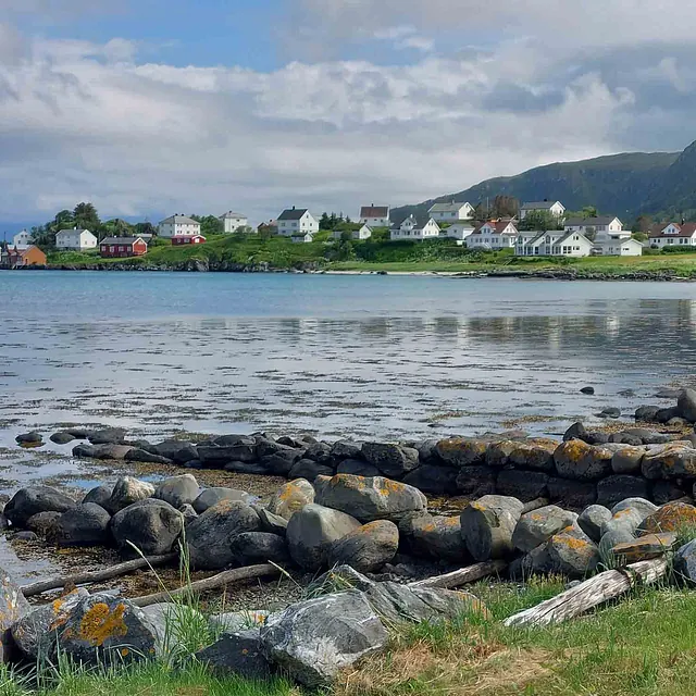 En steinete strandkant i forgrunnen med utsikt over en fjord mot en liten landsby med hvitmalte hus.