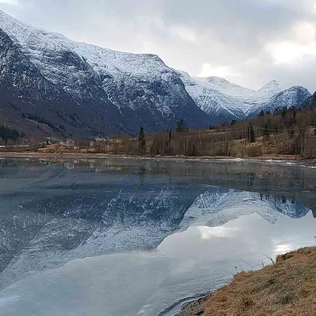 Delvis frosset eller blikkstille vann som speiler snødekte fjelltopper under en vinterhimmel.