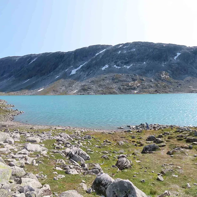 Turkis innsjø omkranset av steinete terreng og høye fjell under en blå himmel.