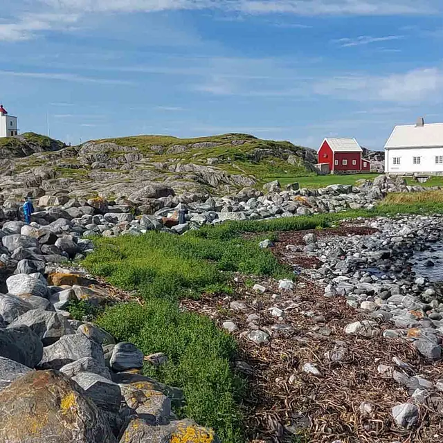 Svaberg og rullesteiner i forgrunnen med et hvitmalt hus og et lite sjømerke under en blå himmel.