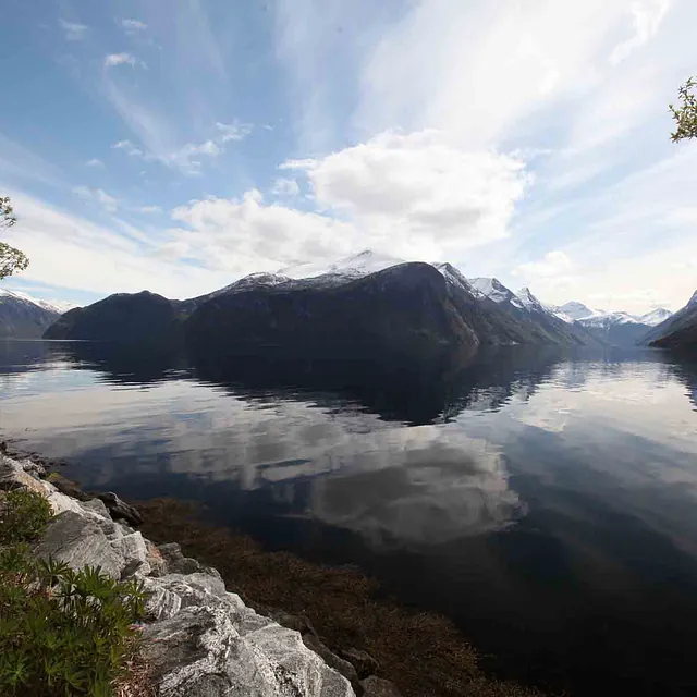 Vidstrakt panoramautsikt over en rolig, mørk fjord som reflekterer en skyet himmel og fjellsider.