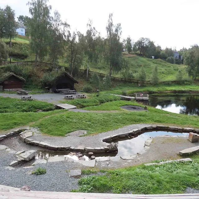 Gammelt gårdstun med tradisjonelle torvtekte tak, gressplen og steinmurer omkranset av skog.