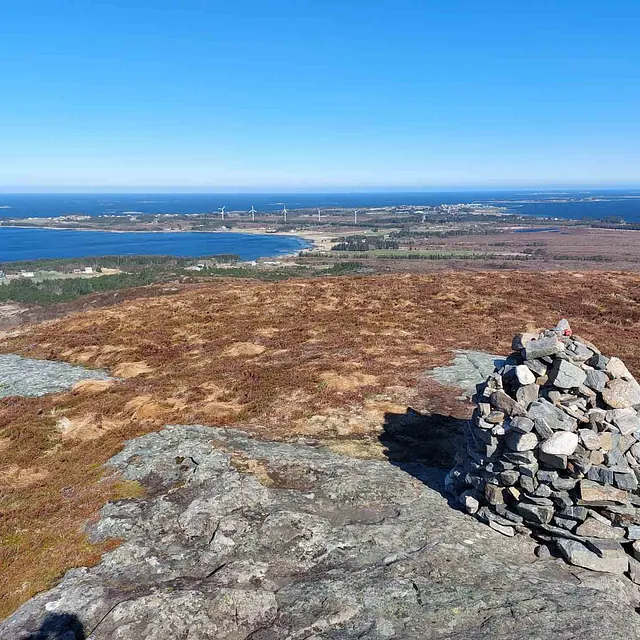 Luftig utsikt fra en fjelltopp med en steinvarde, over et flatt kystlandskap som strekker seg mot havet.