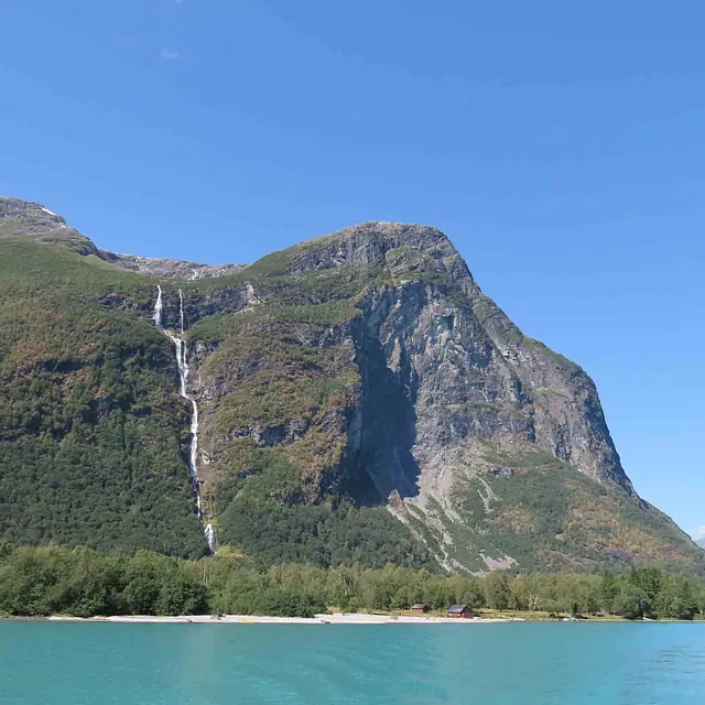 Høyt, bratt fjell med en tynn foss som renner ned mot et turkisblått vann.
