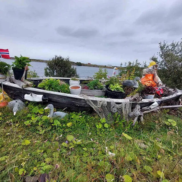 En gammel trebåt dekorert med blomsterpotter, figurer og et lite norsk flagg, plassert på en gressplen.